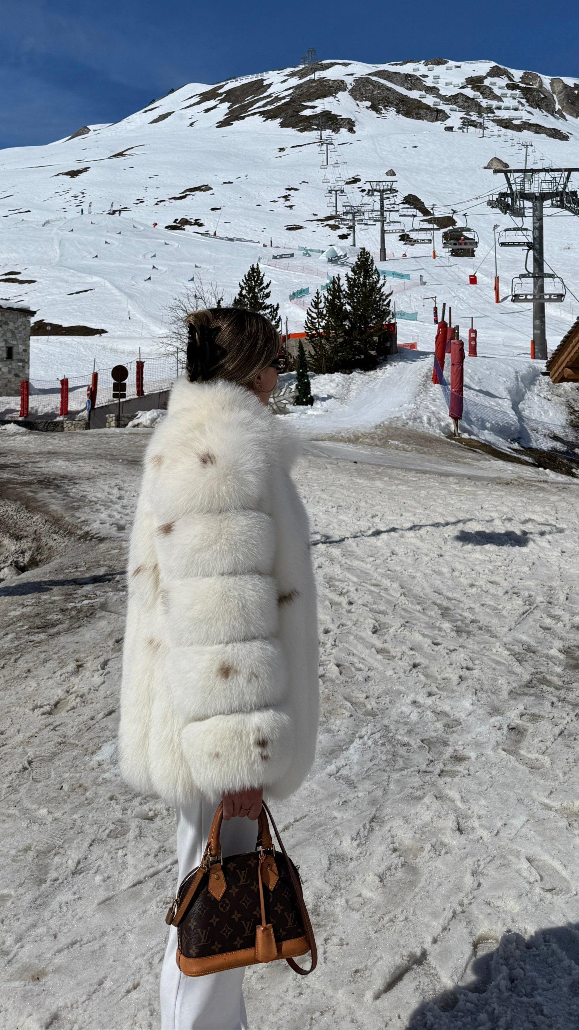 Person in a white fur coat standing on a snowy mountain with ski lifts and mountains in the background.